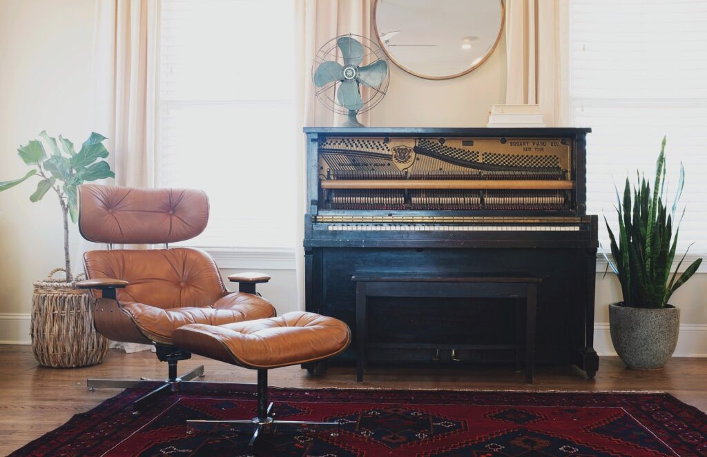 brunt piano i ett rum med vita väggar, trägolv. på golvet ligger en röd matta, det står en beige skinnfåtölj med fotpall bredvid pianot. Bakom fåtöljen står en grön krukväxt i en bambukruka