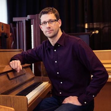 Bild på Fredrik Almeby - man med mörkt hår, glasögon, vinröd skjorta, Han sitter vid ett brunt piano.